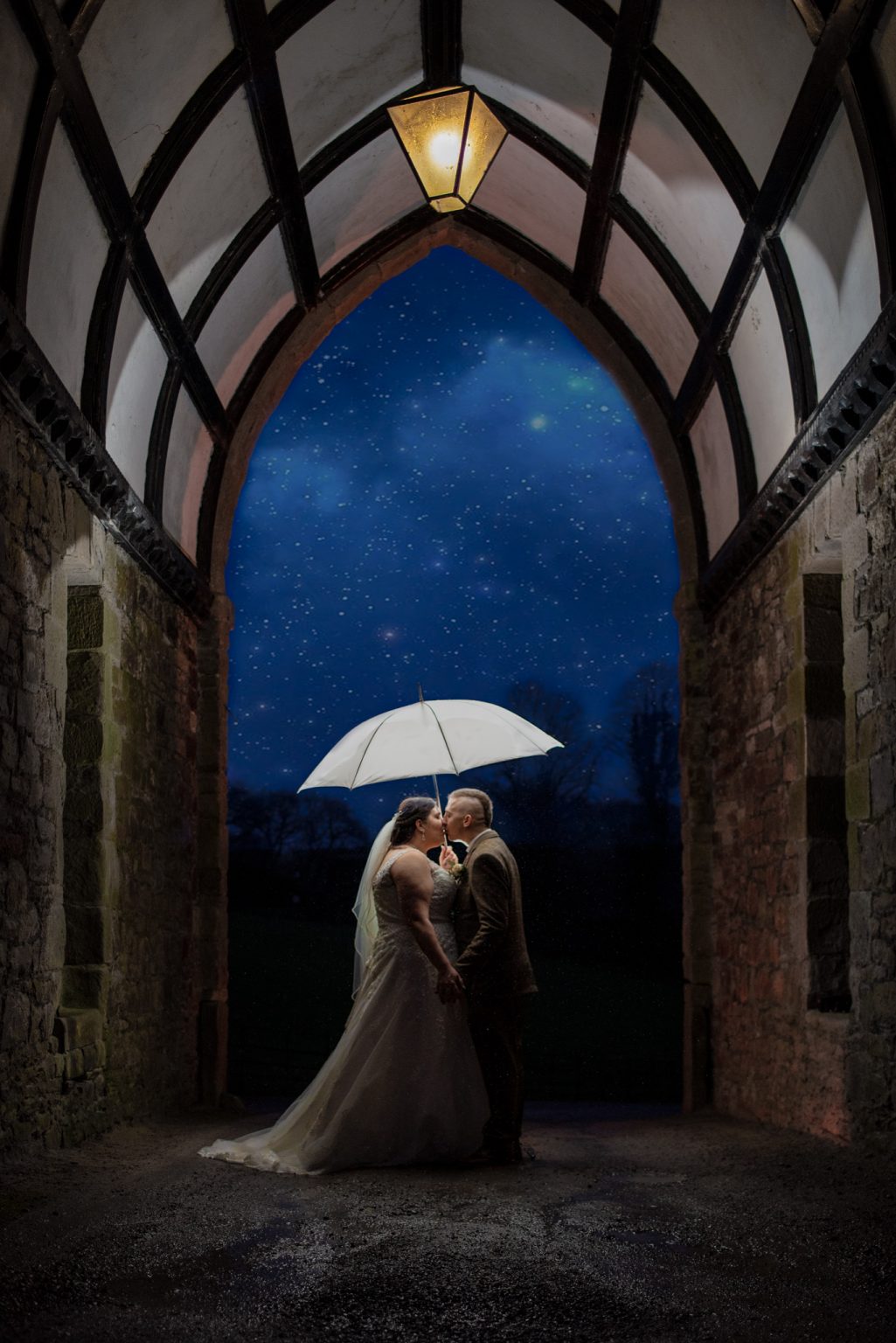 Under the arch shot at Clearwell castle. A flash lights up the archway in the dar as a couple kiss under a white umbrella. Cotswold wedding photography by Pedge Photography