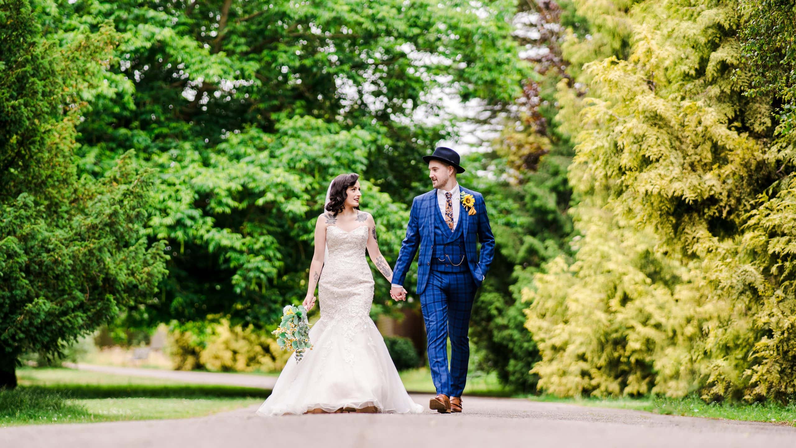 A gorgeous newlywed couple walk through Pittville Park. The bride is in a gorgeous, traditional white dress with an elegant bouquet of sunflowers. The groom is wearing a blue chequered three piece suit, matched with a bowler hat.