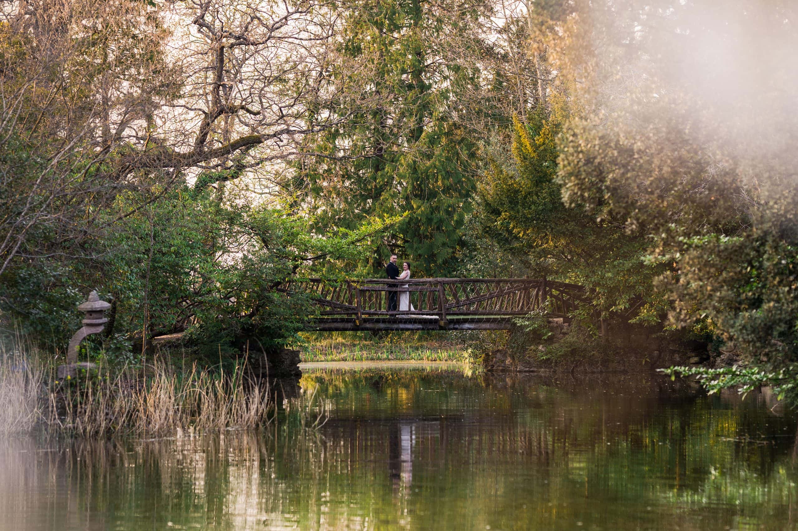 Newlyweds pose on a bridge over looking the water on the grounds of the stunning wedding venue Manor by the lake in Cheltenham, which is one of the top 15 wedding venues in The Cotswolds