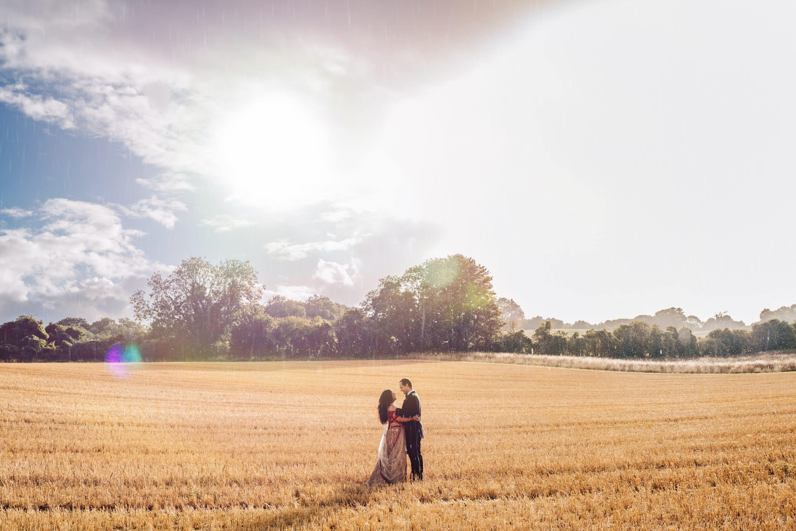 Indian bride at the Barn at Upcote wedding venue stands with her husband in a field of wheat, with the most dramatic scene of sun and rain.