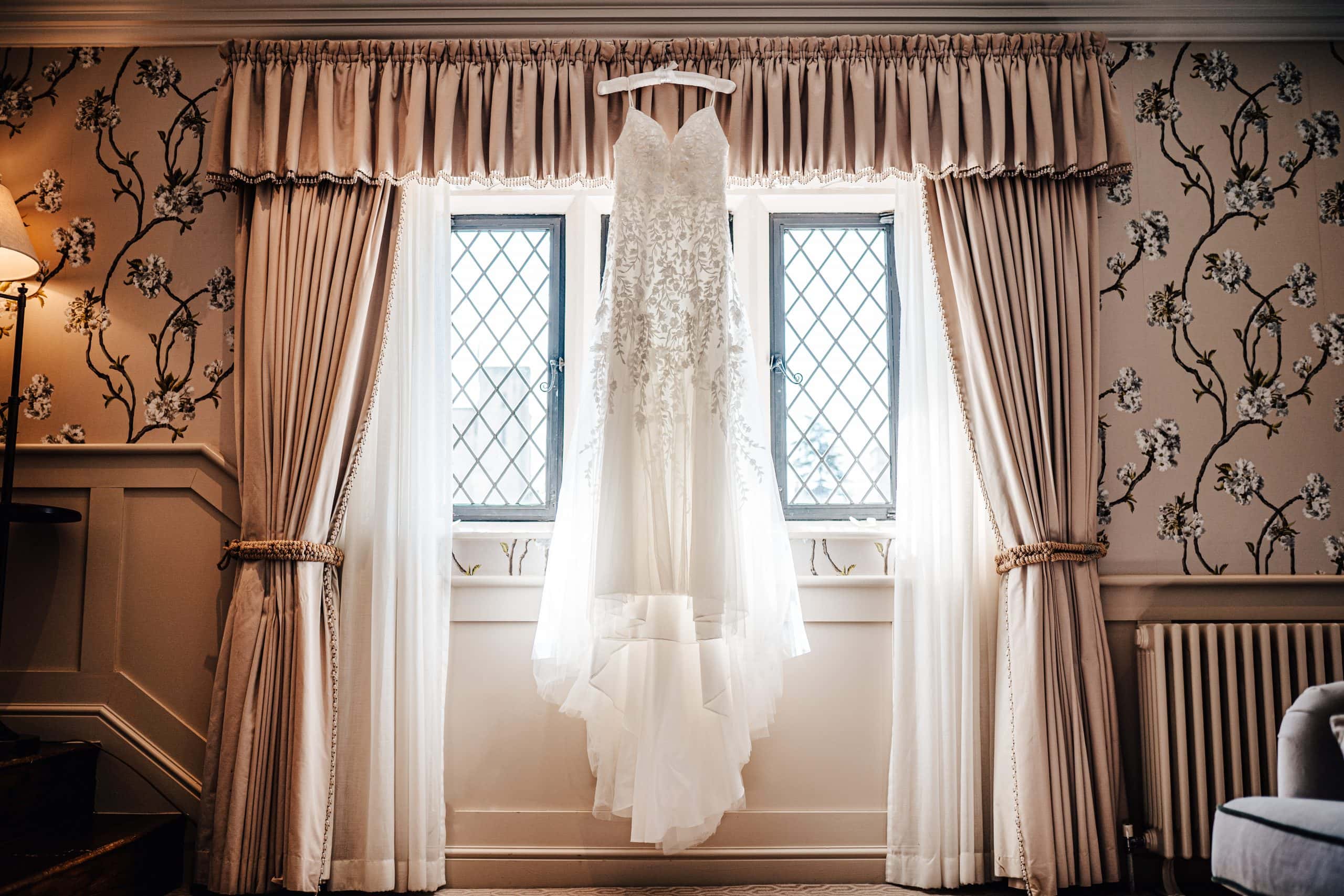 Wedding dress hangs over the curtain rail with the sun pouring through the window at wedding venue Ellenborough Park Hotel